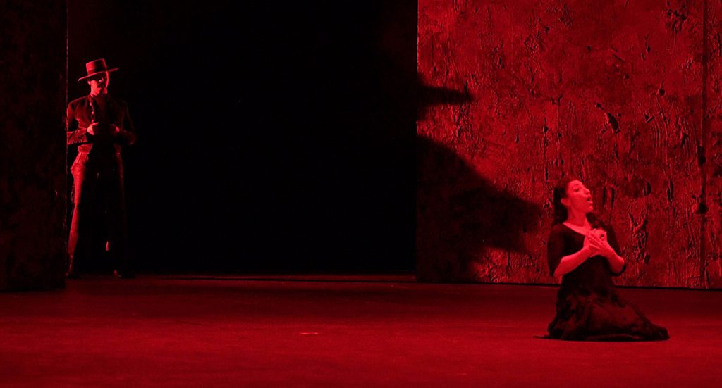 Momento del espectáculo  La vida breve de Manuel de Falla. Imagen con tonos negros y rojos, la protagonista de rodilla y detrás un señor observando | Sevilla Senior
